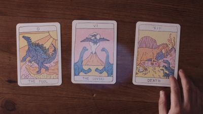 Film still from 'My Diagnosis' (2025) by Jon Beaton. A top-down view of three tarot cards laid on a wooden table. The cards are The Fool, The Lovers and Death. A hand is touching the third card.